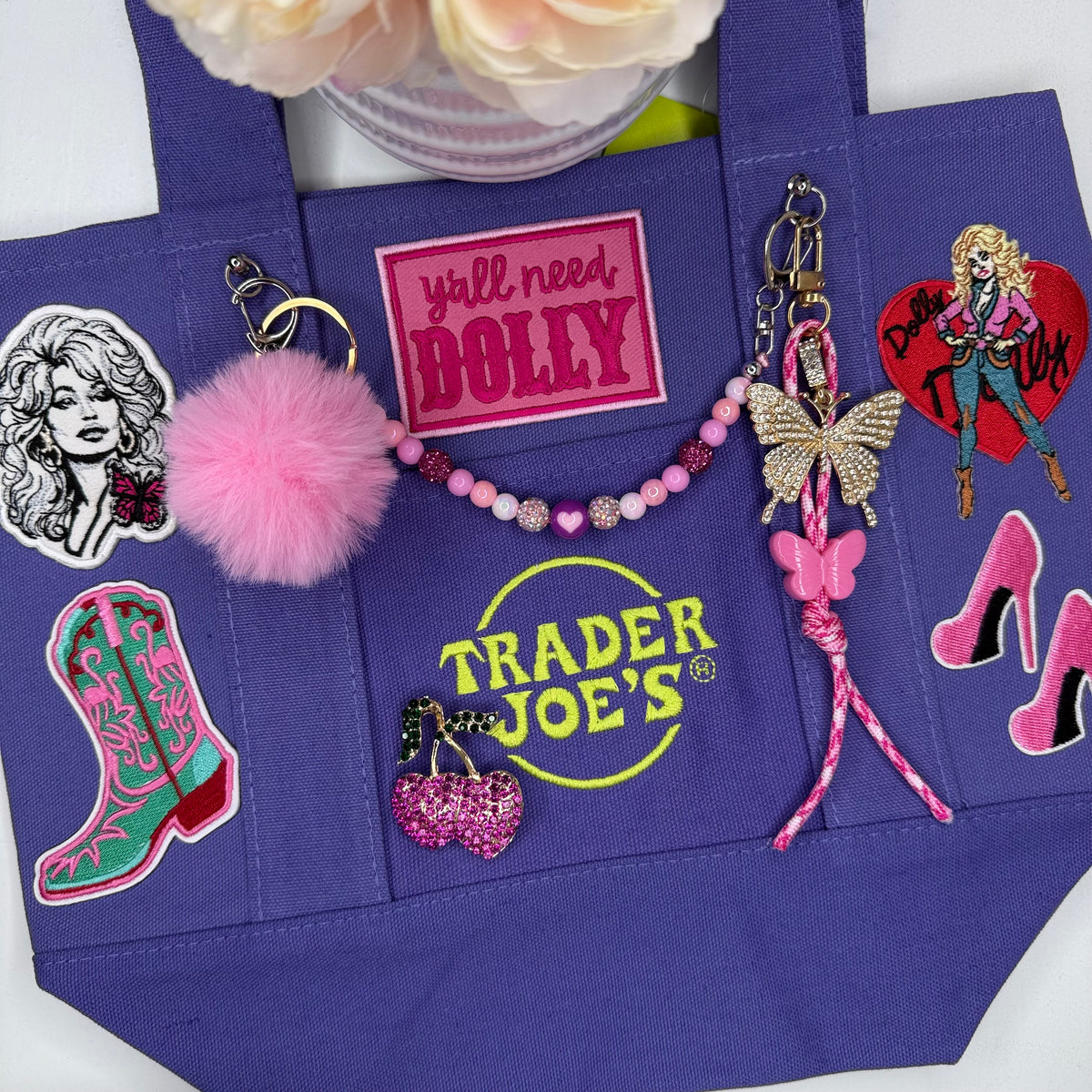 A purple tote bag with embroidered patches and keychain accessories, including a pink pom-pom, beaded necklace, butterfly charm, and cowboy boot patch. Dolly Parton Boots & Heels mini tote by PrettyMUCH.