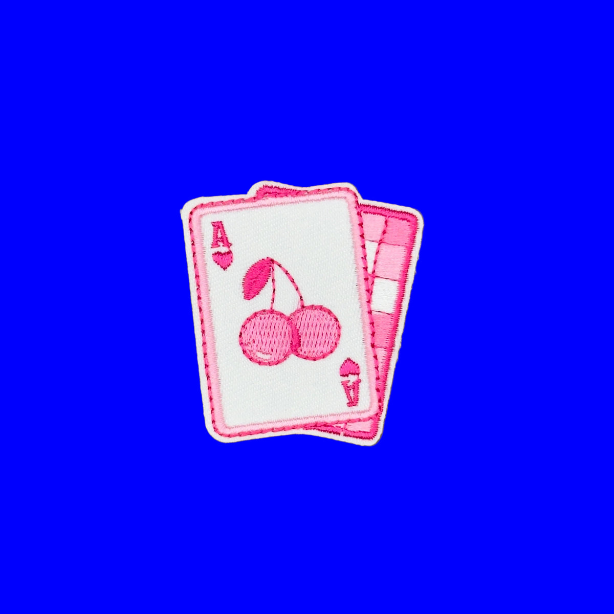 A pink and white embroidered patch shaped like playing cards, featuring two cherries on the front card and a striped pattern on the back.