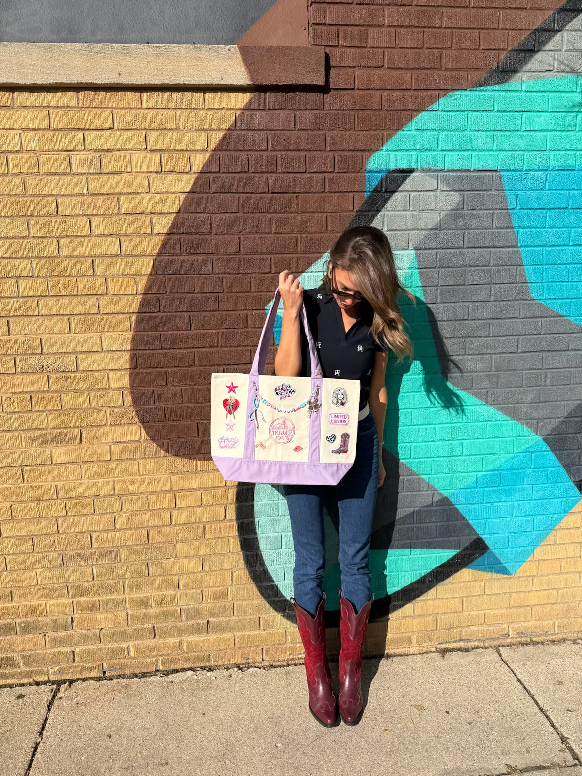 A woman stands outdoors against a painted brick wall holding a lavender tote bag decorated with colorful patchwork designs. She is wearing a dark sleeveless top, blue jeans, and red cowboy boots. Dolly Parton Howdy Darlin' large tote by PrettyMUCH.