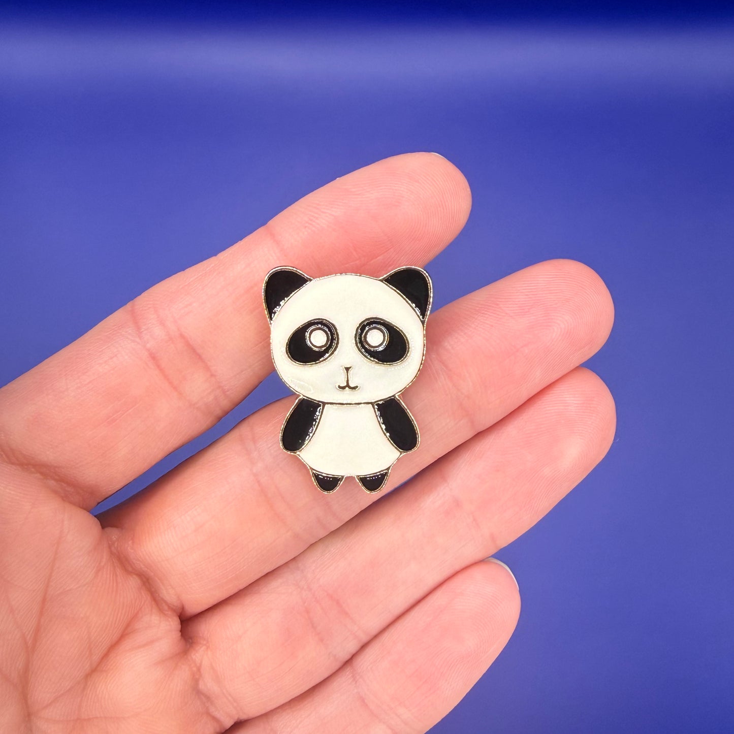 Wide-eyed panda enamel pin with pink cheeks resting on an open palm, blue background