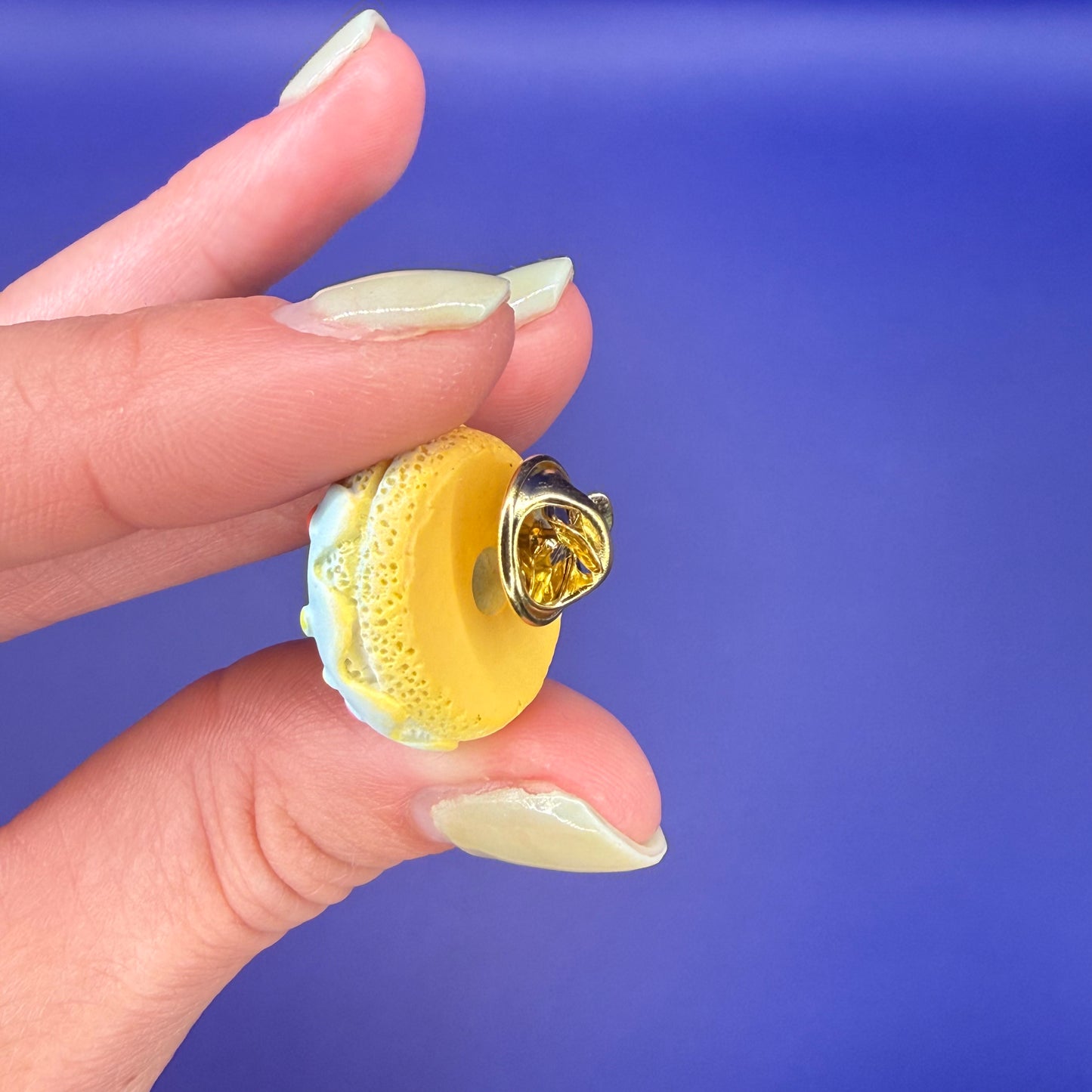Back of the blue daisy sprinkle donut enamel pin showing the gold clutch back, held in hand