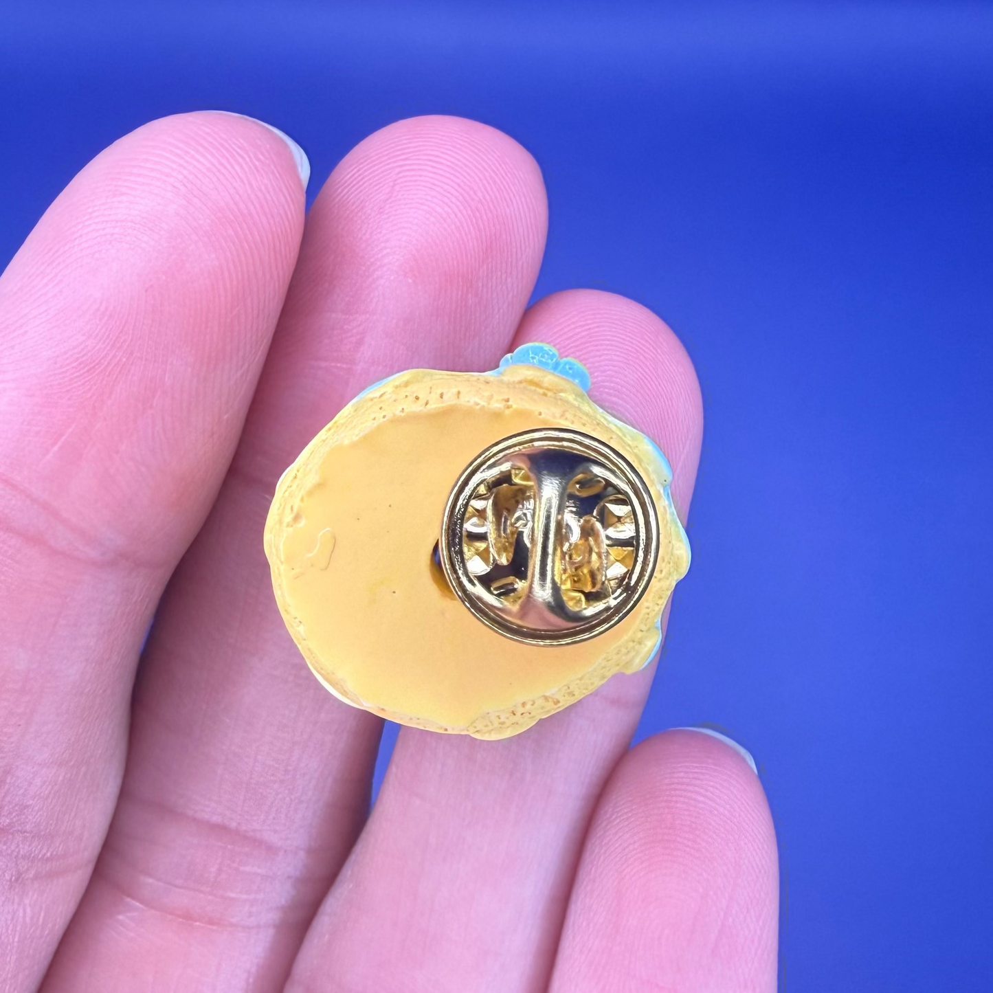 Back of the yellow sprinkle donut enamel pin showing the gold clutch back, held in hand