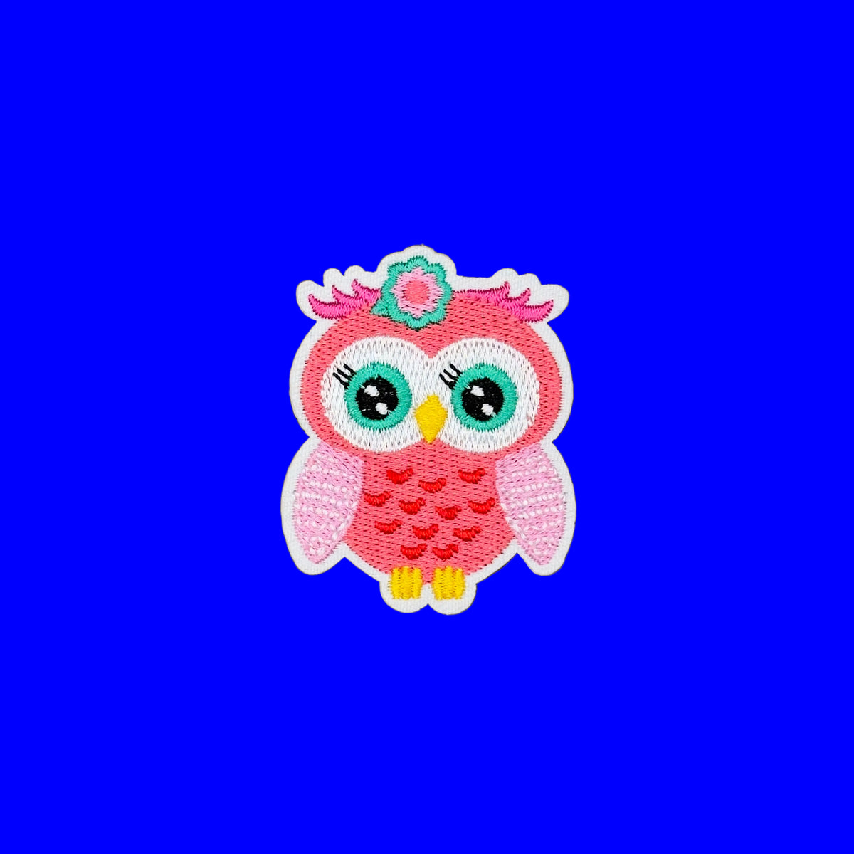 A pink embroidered owl patch with large green eyes, a yellow beak, yellow feet, and a small flower on its head, shown on a plain brown background.