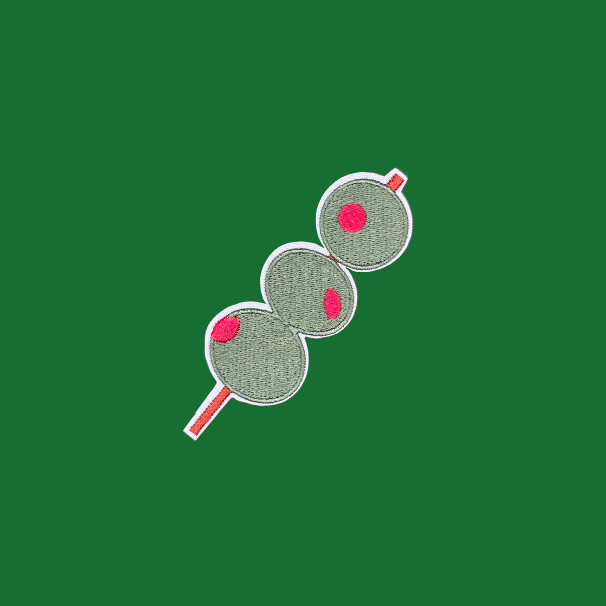 An embroidered patch of three green salami slices on a red skewer, displayed on a solid dusty rose background.