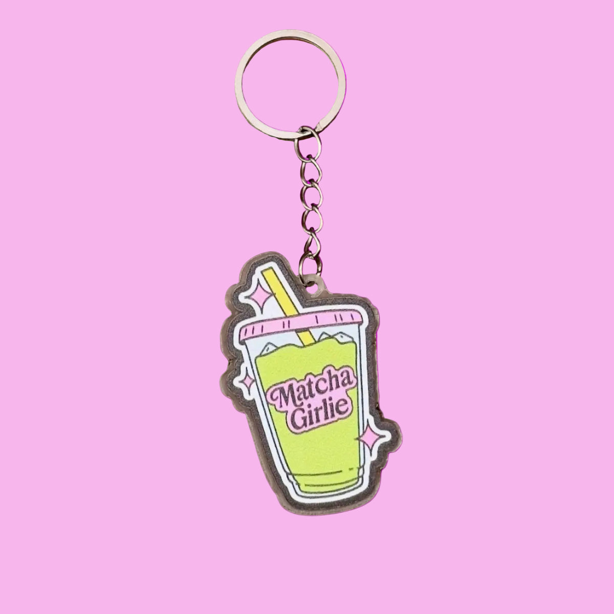 A novelty keychain featuring a cartoon-style matcha green drink cup with the text 'Matcha Girl' in pink, attached to a silver metal key ring and chain, set against a solid pink background.