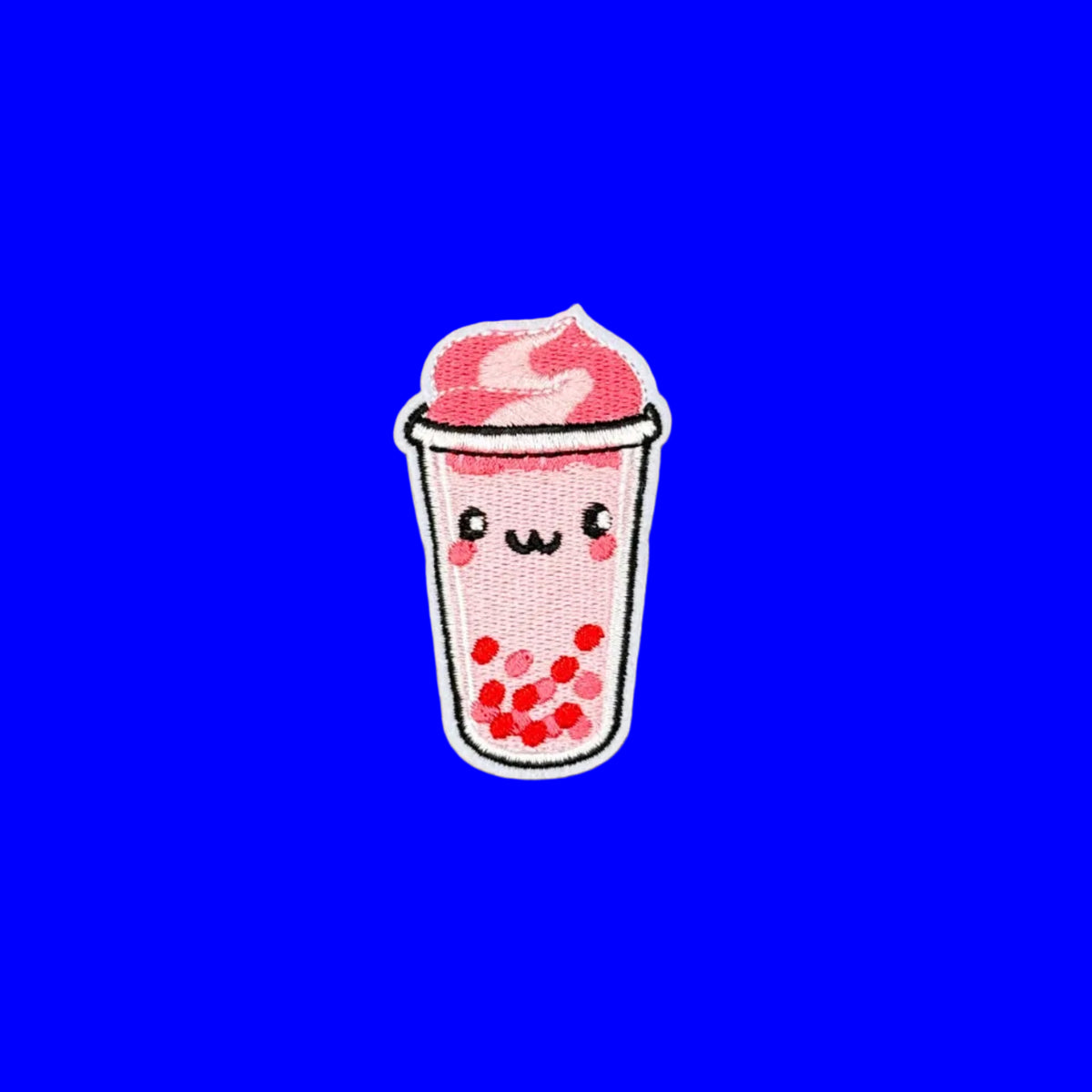 A small embroidered patch of a cute boba drink character, featuring pink foam, red boba pearls, and a smiling face on a white cup, set against a solid brown background.