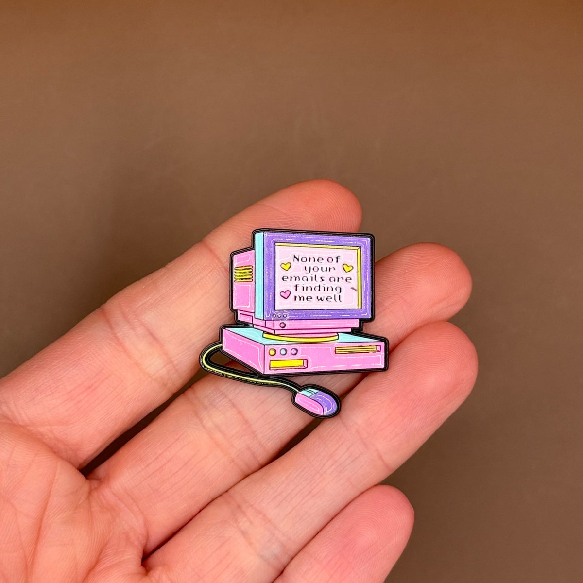 A small enamel pin shaped like a retro computer monitor and keyboard, held on a person's fingers against a plain brown background.