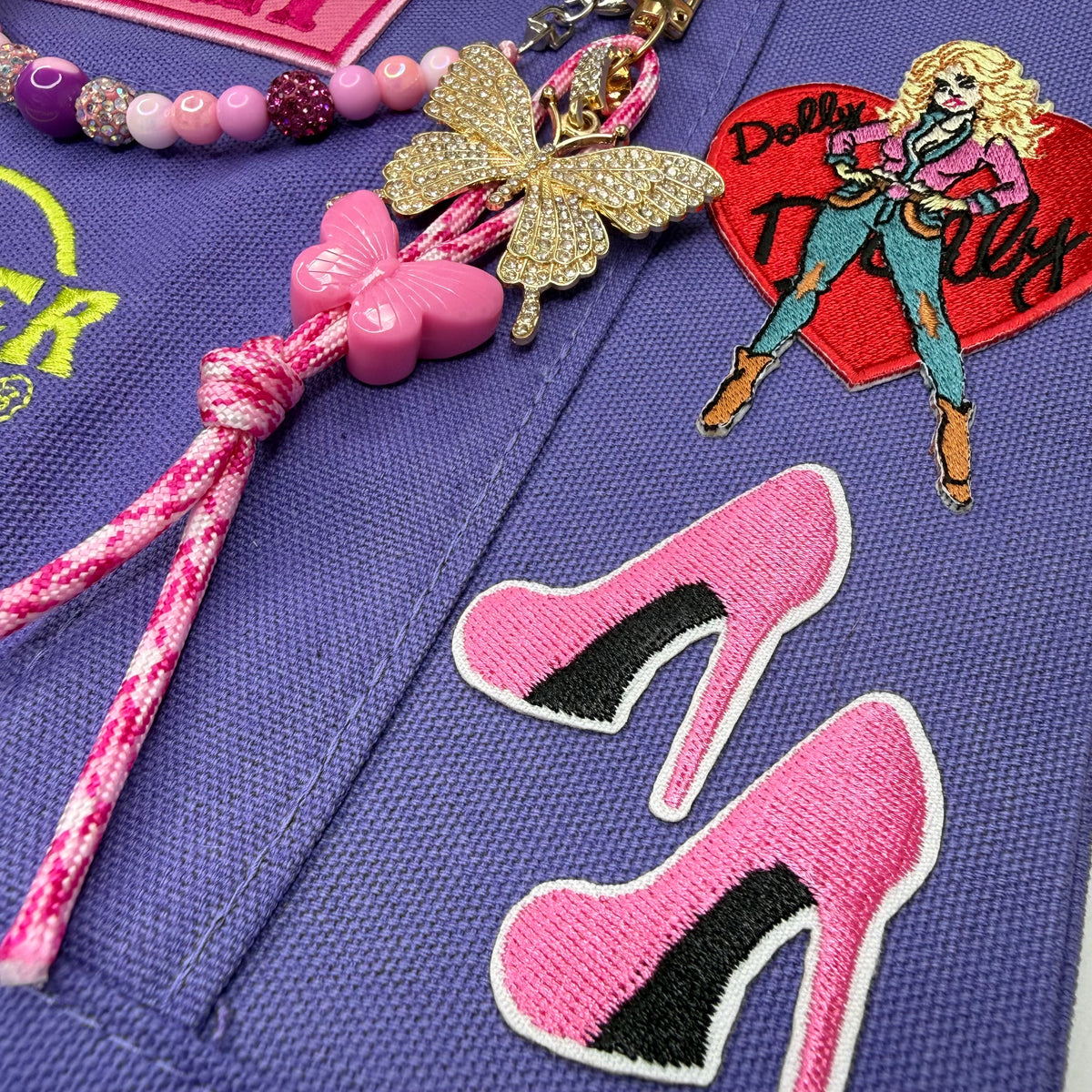 A close-up of a purple woven fabric surface featuring embroidered patches, including pink high-heel shapes, a gold butterfly embellishment, a pink cord tassel, a pink butterfly charm, and a red heart-shaped patch with a stylized woman and the text 'Dolly Parton'. Dolly Parton Boots & Heels mini tote by PrettyMUCH.