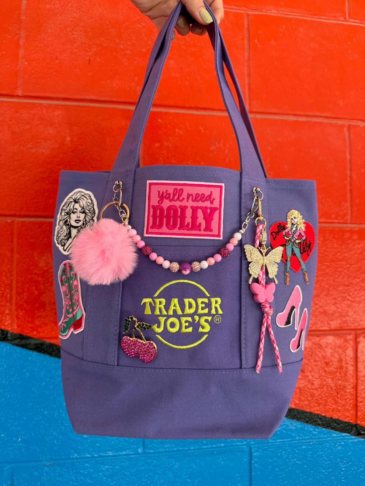 A purple Trader Joe's mini tote decorated with Dolly patches, pink stiletto patches and charms, held up by a hand against a bright orange and blue brick wall. Dolly Parton Boots & Heels mini tote by PrettyMUCH.
