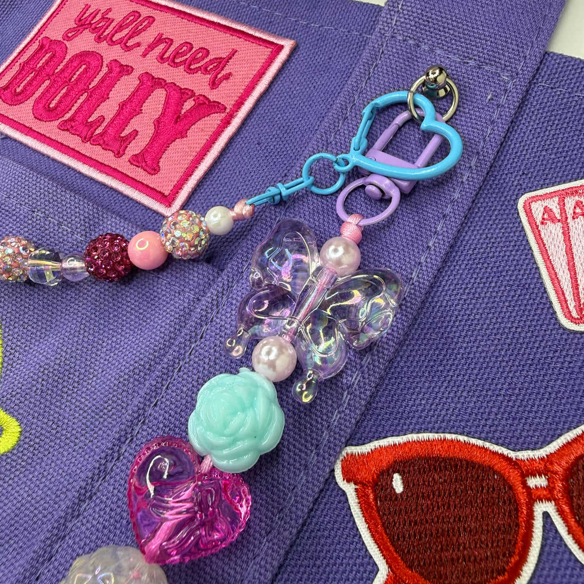 A close-up of a colorful beaded keychain or bag charm attached to a purple fabric strap. The charm includes a light blue heart clasp, a lavender ring, a translucent iridescent butterfly, a pale mint rose, a pink heart, and pink and clear beads. Dolly Parton Aces High mini tote by PrettyMUCH.