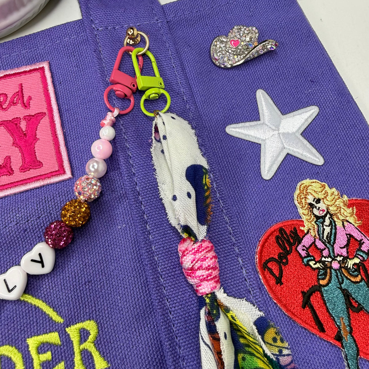 A close-up of a purple embroidered tote bag decorated with patches, pins, and a colorful fabric keychain. The keychain features pink and green clasps, white and pink beads, a glittery gold bead, and a patterned white fabric tassel. Dolly Parton Y'all Need Dolly mini tote by PrettyMUCH.
