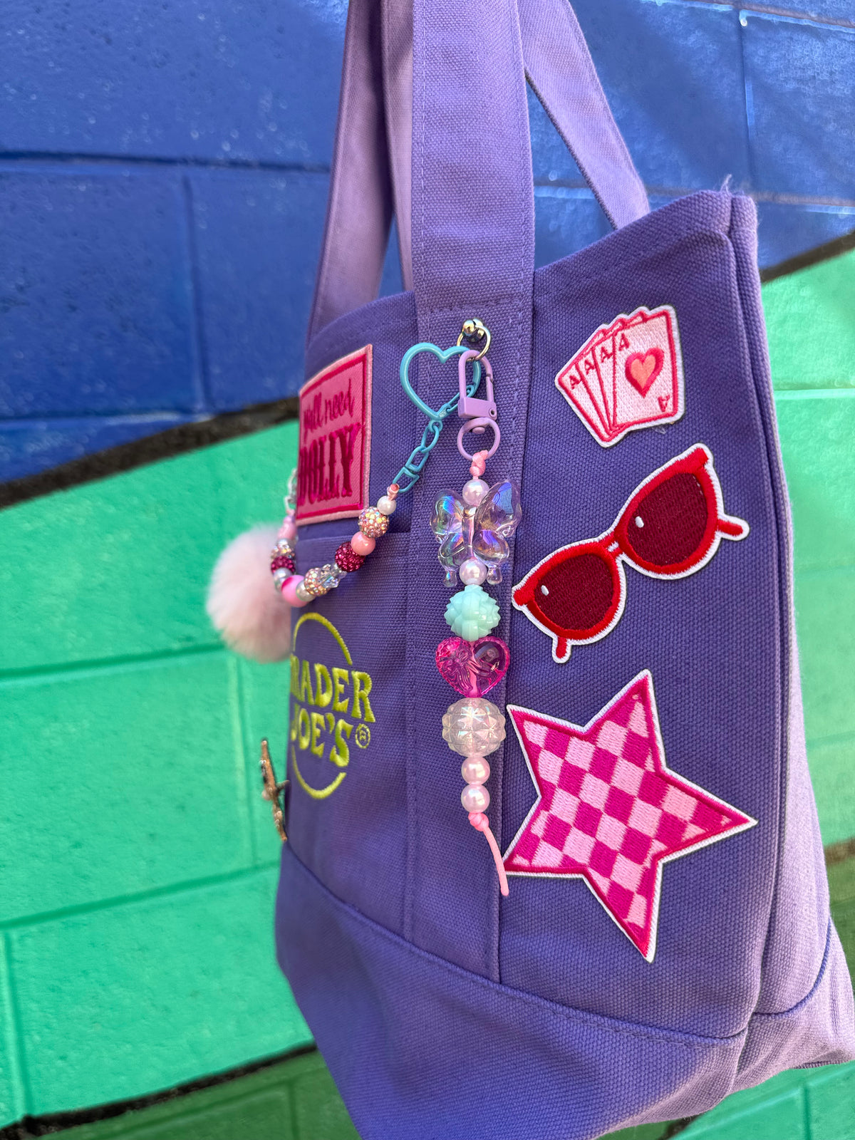 A purple canvas tote bag decorated with colorful patches, including a pink star, red sunglasses, playing cards, and a beaded charm, set against a blue and green painted brick wall. Dolly Parton Aces High mini tote by PrettyMUCH.