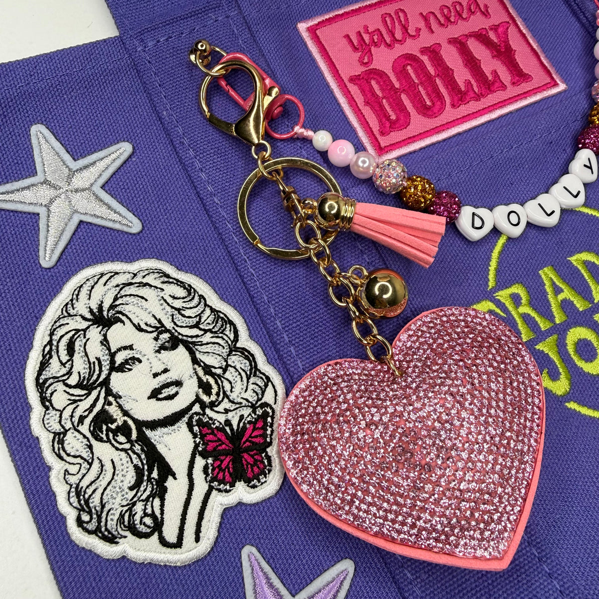 A close-up of a pink rhinestone-embellished heart-shaped keychain charm with a pink tassel and gold-tone keyring, laid on a purple embroidered fabric surface with decorative patches and beads. Dolly Parton Y'all Need Dolly mini tote by PrettyMUCH.