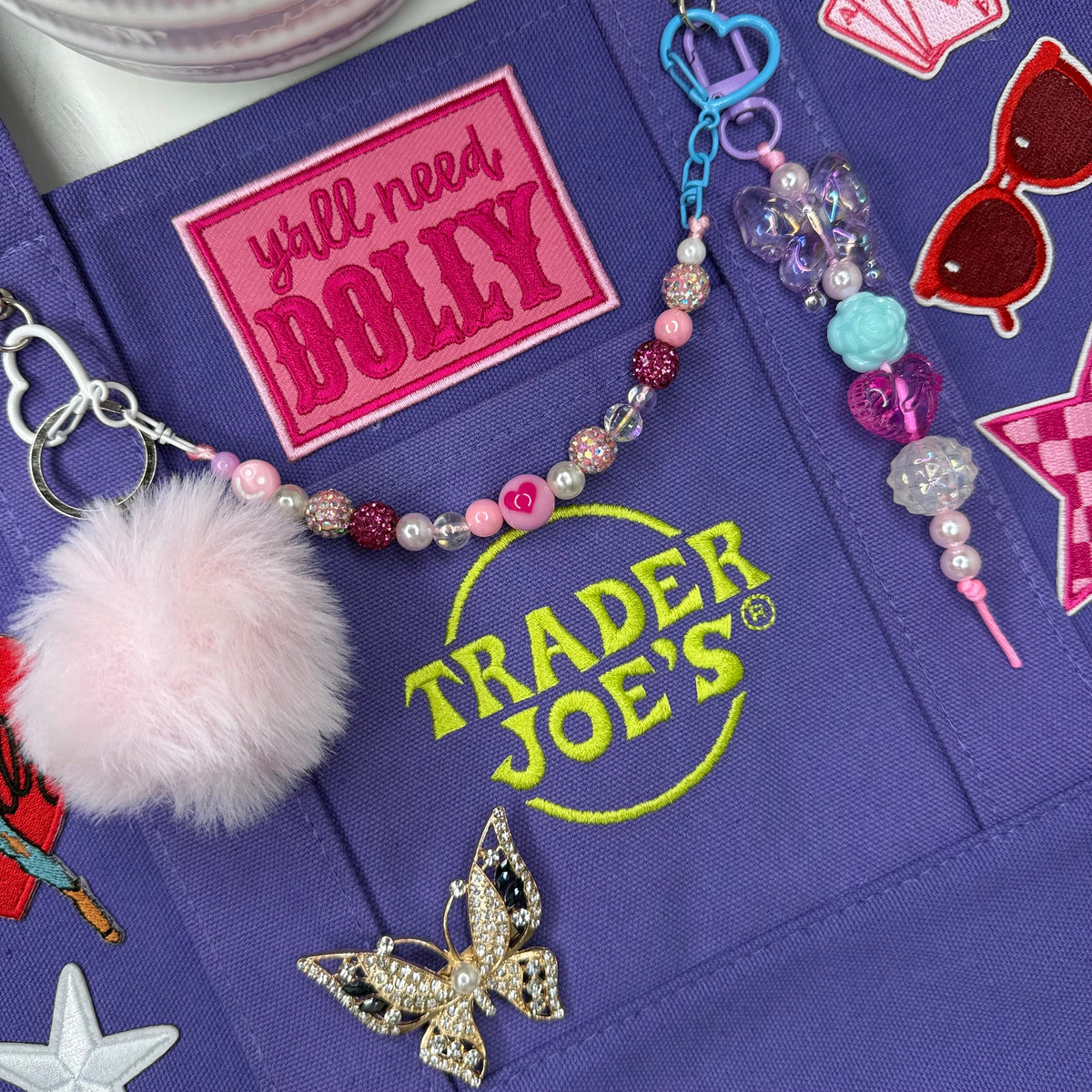 Close-up of the purple Trader Joe's mini tote with the hot pink 'y'all need DOLLY' patch, the bright green embroidered Trader Joe's logo and a beaded butterfly charm with a fluffy pink pom-pom keychain. Dolly Parton Aces High mini tote by PrettyMUCH.