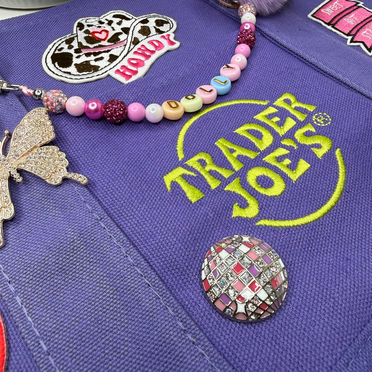 Close-up of the purple Trader Joe's mini tote with the yellow embroidered Trader Joe's logo, a cow-print HOWDY cowgirl hat patch, a beaded DOLLY bag chain, a rhinestone butterfly charm and a pink-and-purple rhinestone disco ball pin. Dolly Parton Sweet but Strong mini tote by PrettyMUCH.