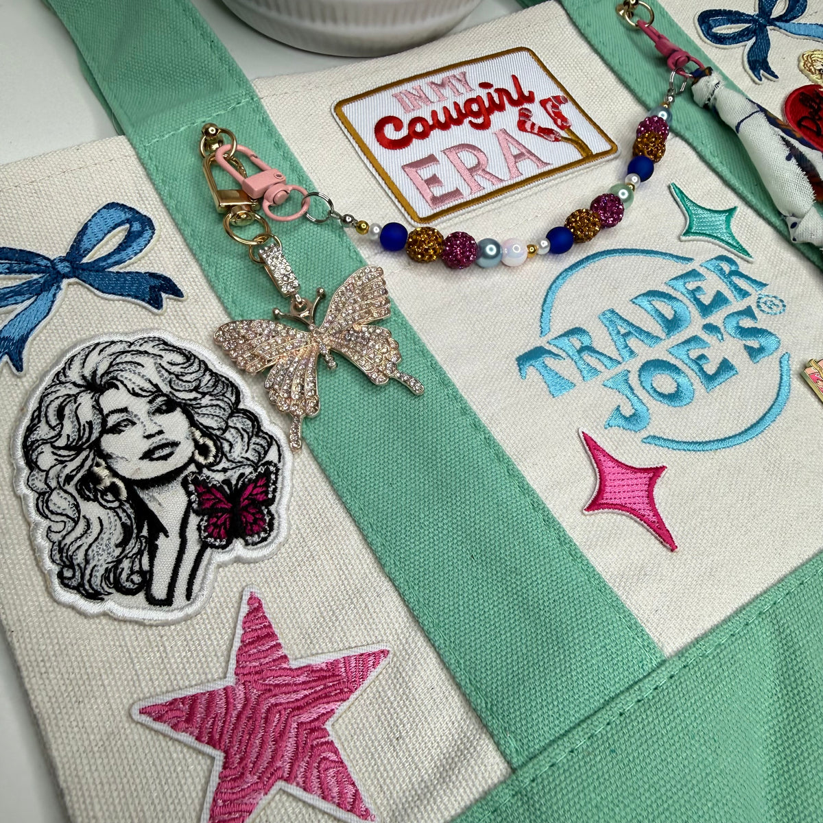 A close-up of a canvas tote bag with mint green straps and embroidered details, including a Trader Joe's logo, a pink star, a cowgirl portrait patch, and a beaded keychain with a rhinestone butterfly charm. Dolly Parton Cowgirl Era mini tote by PrettyMUCH.