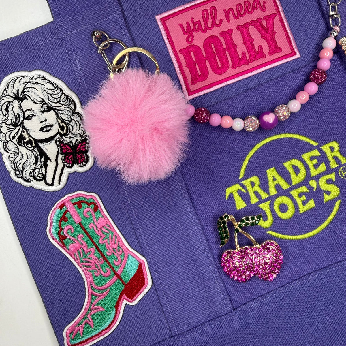 A close-up of a purple fabric tote bag decorated with multiple colorful patches and accessories, including a pink pom-pom keychain, a beaded necklace, a cowboy boot patch, a Dolly patch, and a Trader Joe's patch. Dolly Parton Boots & Heels mini tote by PrettyMUCH.