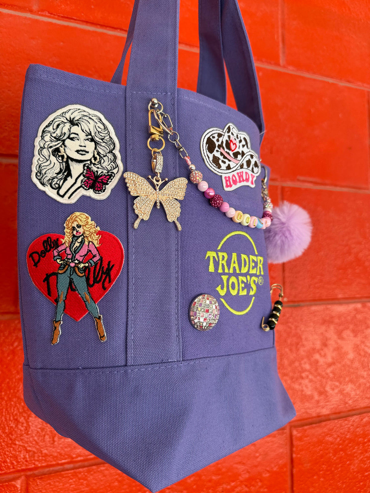 A purple Trader Joe's mini tote decorated with pins, charms and patches, including a gold rhinestone butterfly charm, a lavender pom-pom and the embroidered Trader Joe's logo, set against a bright orange brick wall. Dolly Parton Sweet but Strong mini tote by PrettyMUCH.