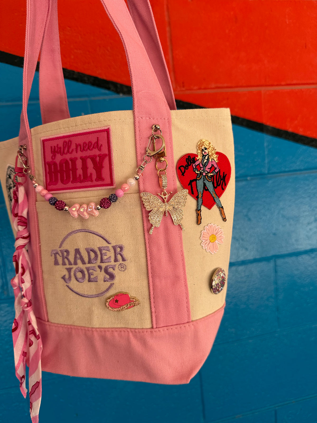 A pink and cream Trader Joe's mini tote decorated with pins, charms and a beaded bag chain, featuring the Trader Joe's logo and the hot pink 'y'all need DOLLY' patch, set against a colorful red and blue background. Dolly Parton Pink Bandana mini tote by PrettyMUCH.
