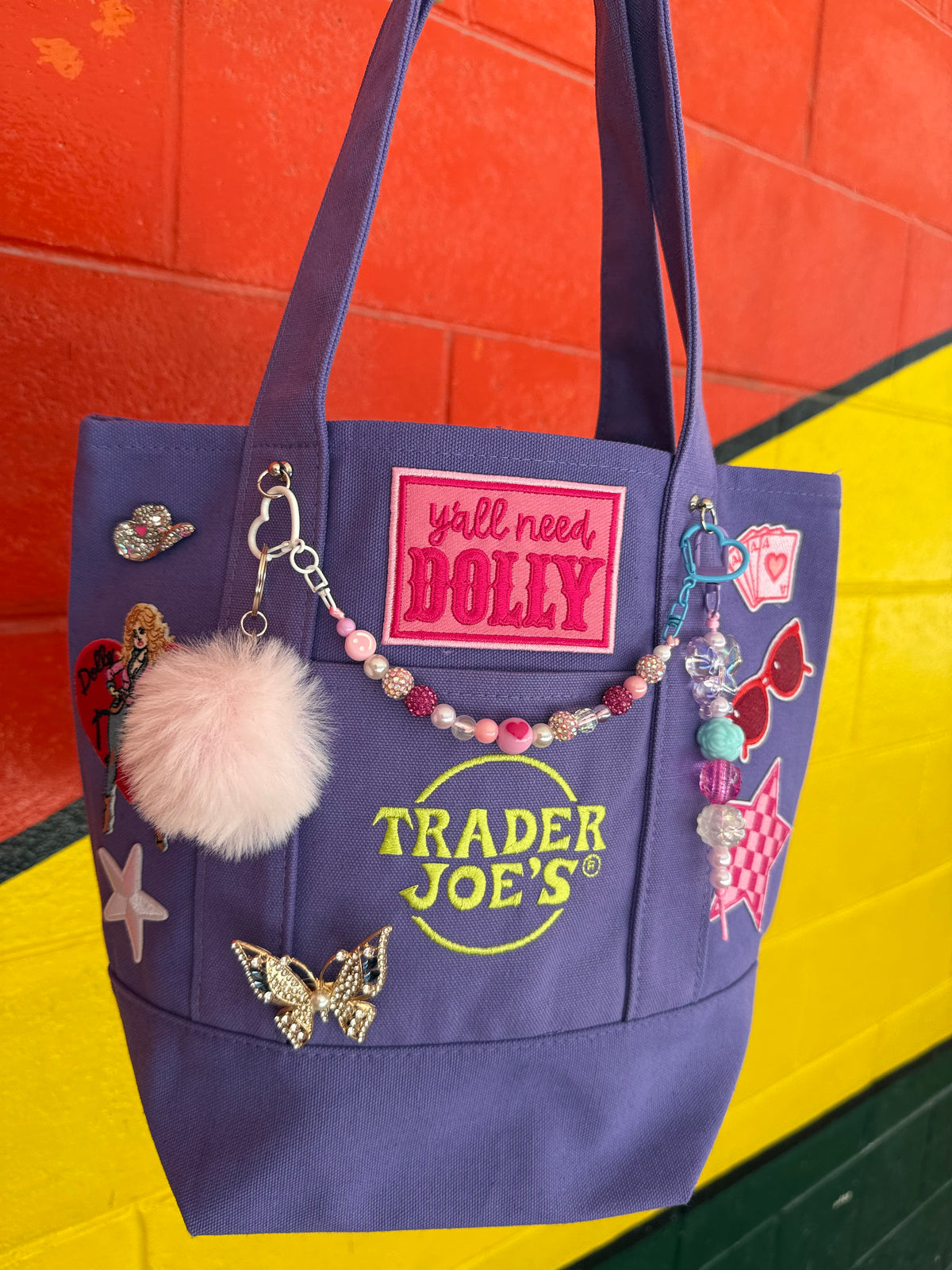 A purple Trader Joe's mini tote with a hot pink 'y'all need DOLLY' patch, the embroidered Trader Joe's logo, a fluffy pink pom-pom and a beaded bag chain, set against a bright orange and yellow brick wall. Dolly Parton Aces High mini tote by PrettyMUCH.