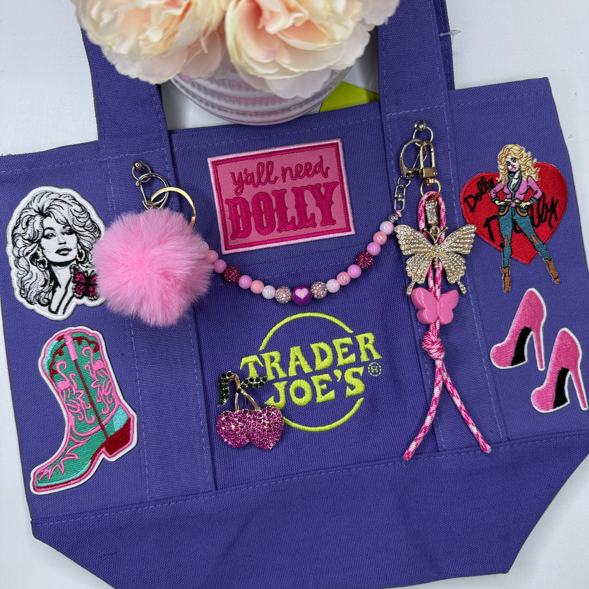 A purple Trader Joe's mini tote decorated with Dolly patches and keychains, including a pink pom-pom, a pink beaded bag chain, a butterfly keychain, the hot pink 'y'all need DOLLY' patch and pink stiletto patches. Dolly Parton Boots & Heels mini tote by PrettyMUCH.