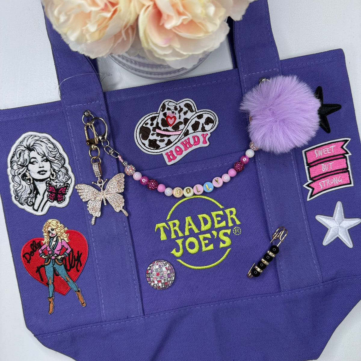 A purple Trader Joe's mini tote decorated with Dolly patches, a lavender pom-pom, a beaded DOLLY bag chain with a rhinestone butterfly keychain and black and white star patches, styled on a white surface with a peach flower in the corner. Dolly Parton Sweet but Strong mini tote by PrettyMUCH.
