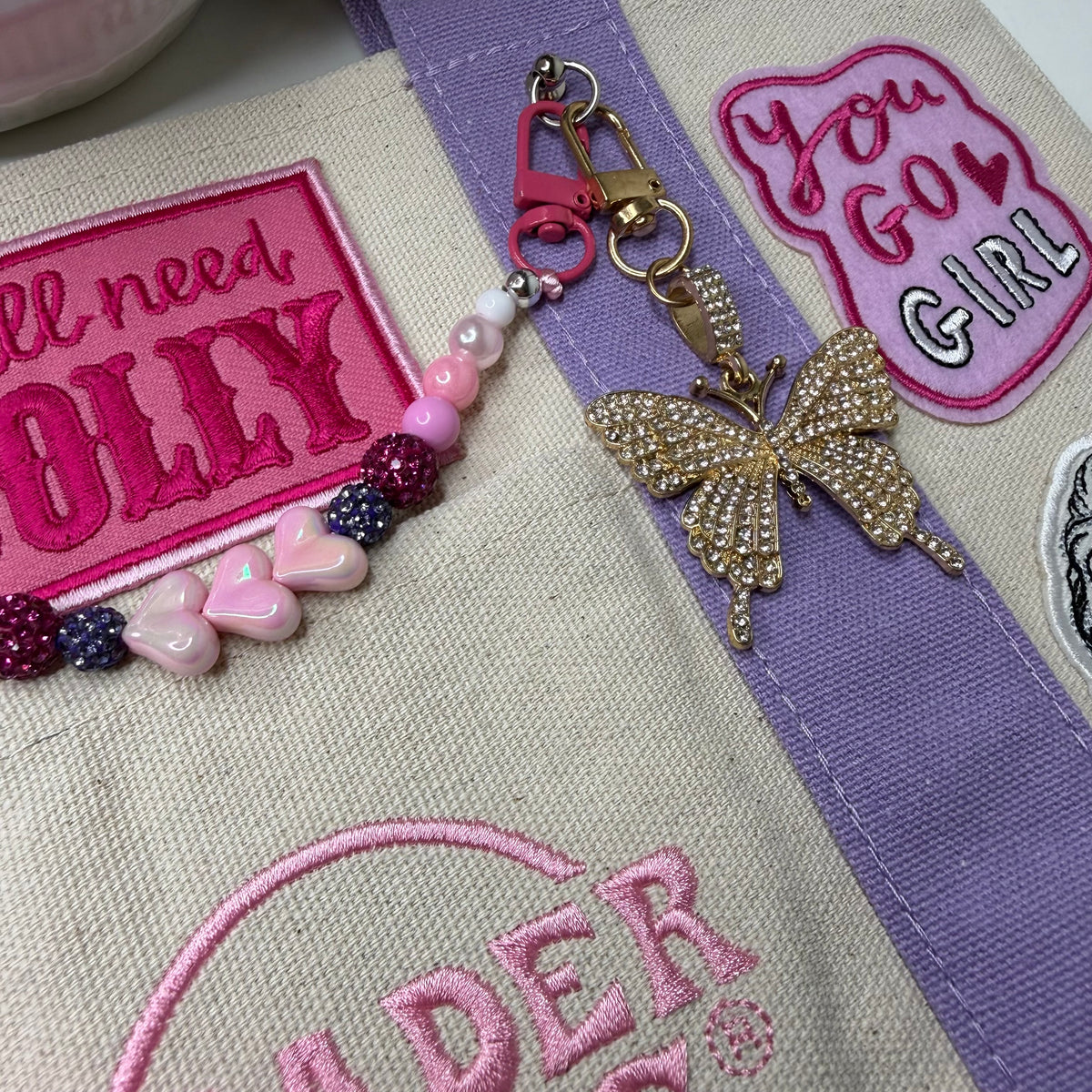 Close-up of the lavender and cream Trader Joe's mini tote with pink embroidered patches and a gold rhinestone butterfly keychain attached near the top. Dolly Parton You Go Girl mini tote by PrettyMUCH.