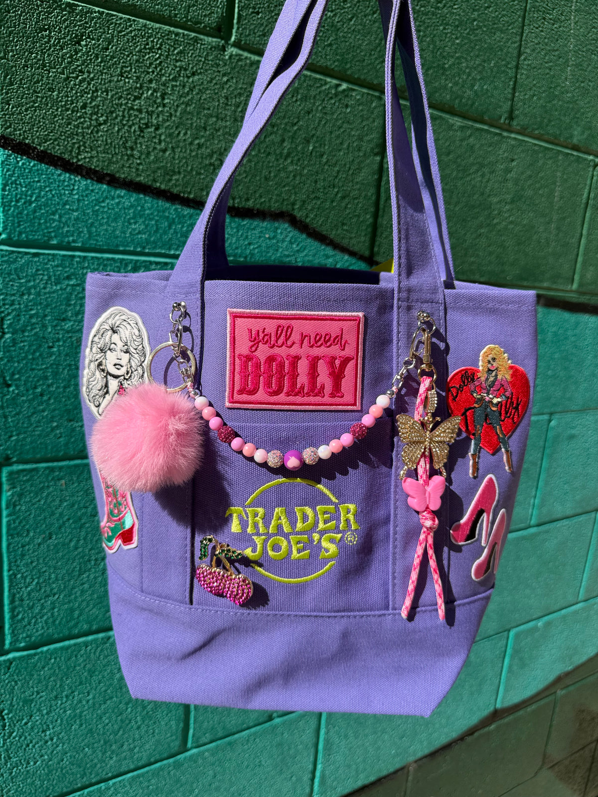 A lavender purple canvas tote bag decorated with various patches, a pink pom-pom, a pink beaded keychain, and a butterfly charm, hanging against a green brick wall. Dolly Parton Boots & Heels mini tote by PrettyMUCH.