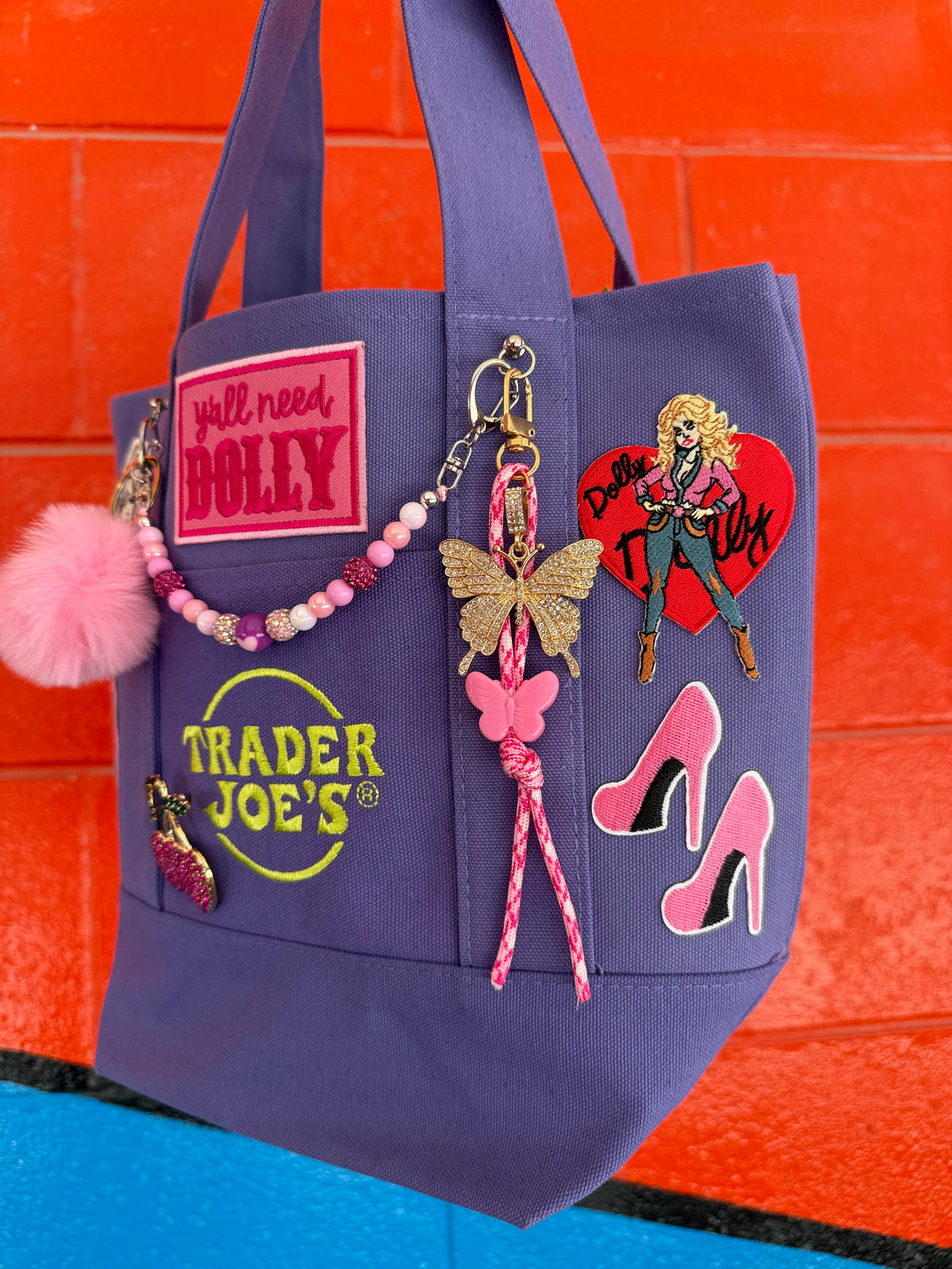 A purple canvas tote bag with multiple decorative patches, charms, and pins, including a pink pom-pom, a Trader Joe's logo patch, and a pink high-heel patch, set against a bright orange brick wall. Dolly Parton Boots & Heels mini tote by PrettyMUCH.