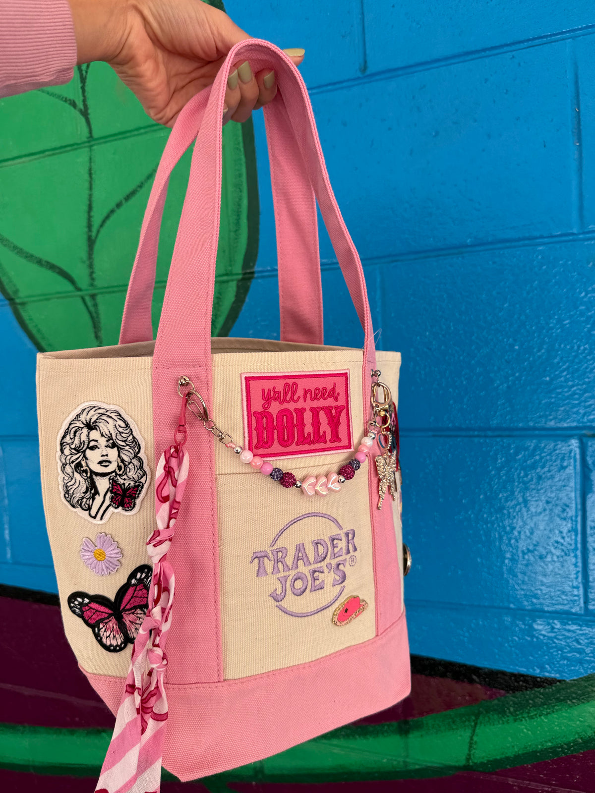 A hand holds a pink and cream Trader Joe's mini tote decorated with Dolly patches, a pink beaded bag chain and a pink bandana, with a hot pink 'y'all need DOLLY' patch alongside the Trader Joe's logo. Dolly Parton Pink Bandana mini tote by PrettyMUCH.
