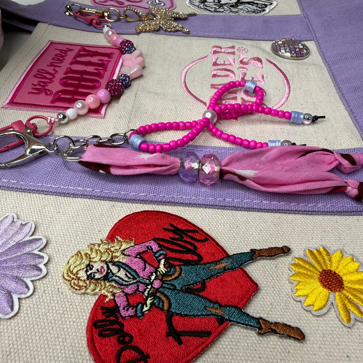 Close-up of the lavender and cream Trader Joe's mini tote with pink and purple embroidered patches, a beaded bag chain and a pink bandana twilly. Dolly Parton You Go Girl mini tote by PrettyMUCH.