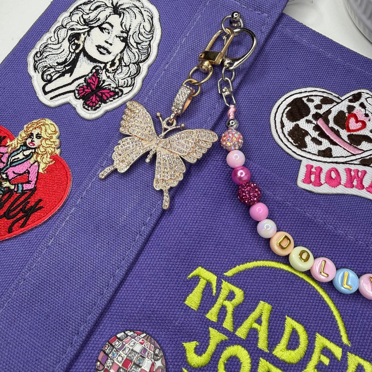 A close-up of a purple embroidered fabric accessory decorated with colorful patches and a rhinestone butterfly charm attached to a beaded strap. Dolly Parton Sweet but Strong mini tote by PrettyMUCH.