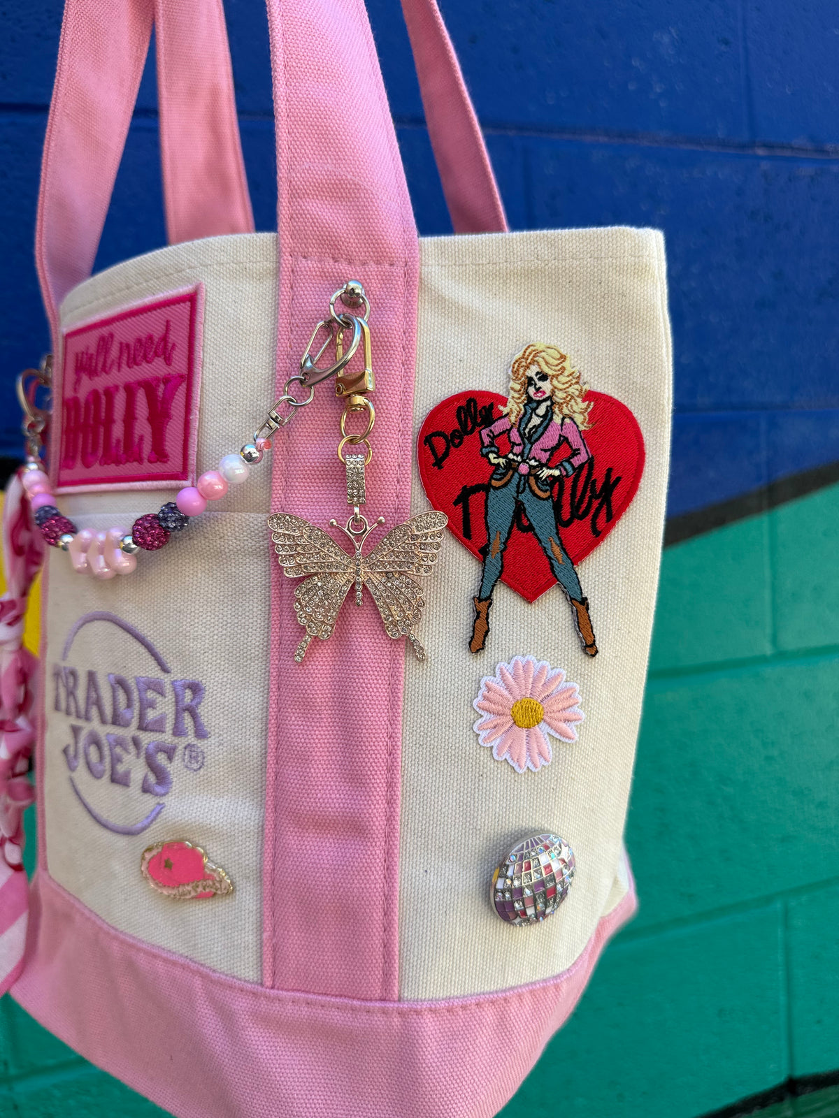 A pink and cream canvas tote bag decorated with various patches, pins, and charms, including a Trader Joe's patch, a butterfly charm, and a pink heart patch, set against a blue and green brick wall background. Dolly Parton Pink Bandana mini tote by PrettyMUCH.