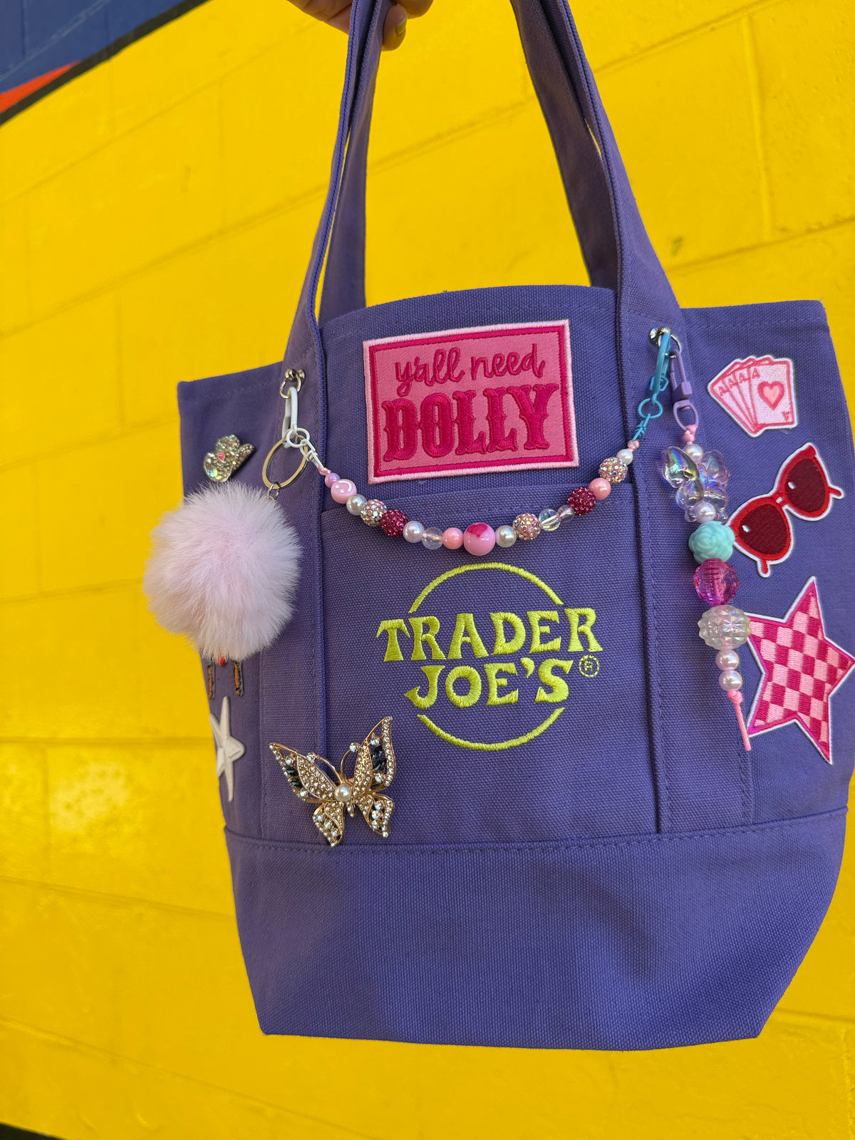 A purple Trader Joe's tote bag decorated with colorful patches, a beaded charm, a pink pom-pom, and a butterfly embellishment, set against a bright yellow brick wall. Dolly Parton Aces High mini tote by PrettyMUCH.