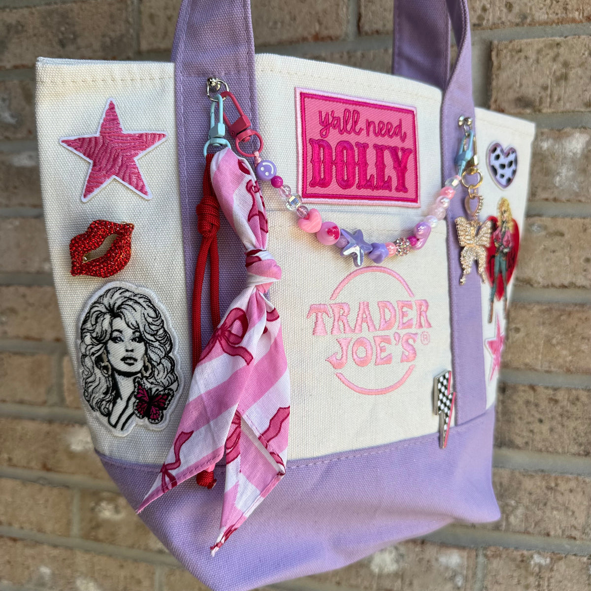 A lavender and cream Trader Joe's mini canvas tote decorated with Dolly patches, a pink striped bandana twilly and a beaded bag chain, hanging against a brick wall. Dolly Parton Pretty in Pastel mini tote by PrettyMUCH.