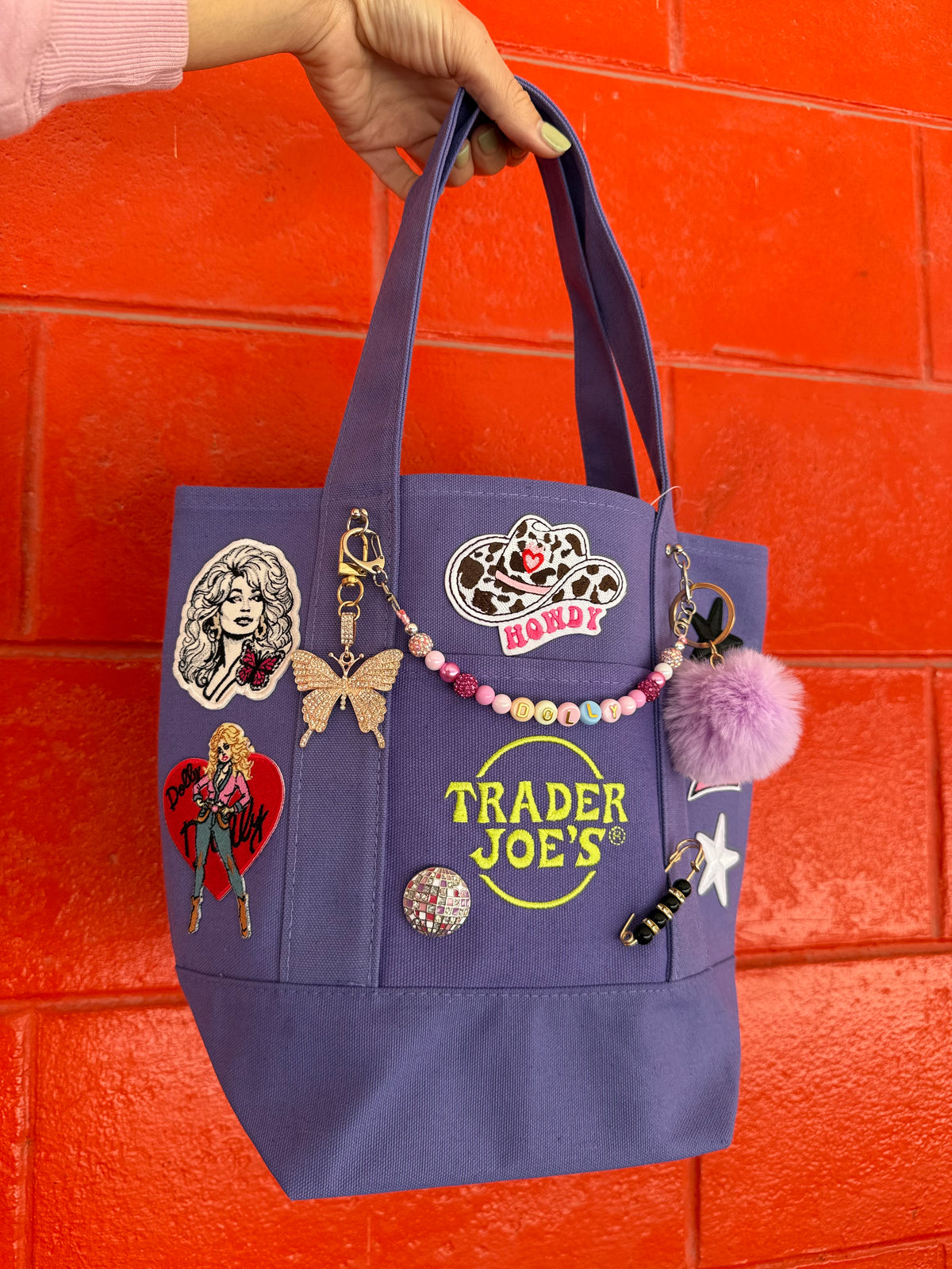 A purple canvas tote bag decorated with various pins, charms, and a beaded keychain, held by a hand against a bright orange brick wall. The bag features the Trader Joe's logo embroidered in yellow on the front. Dolly Parton Sweet but Strong mini tote by PrettyMUCH.