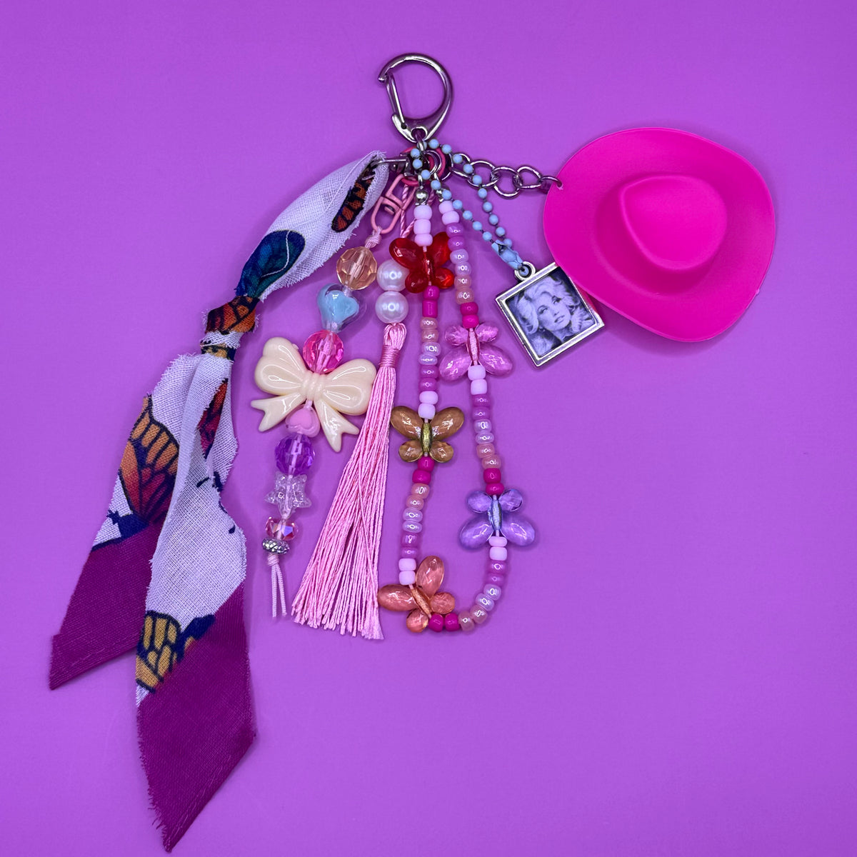 1-of-1 handmade Dolly Parton bag charm with a hot pink cowboy hat, framed Dolly portrait charm, butterfly-print scarf tie, pink silk tassel and beaded butterfly strands on a silver clasp.