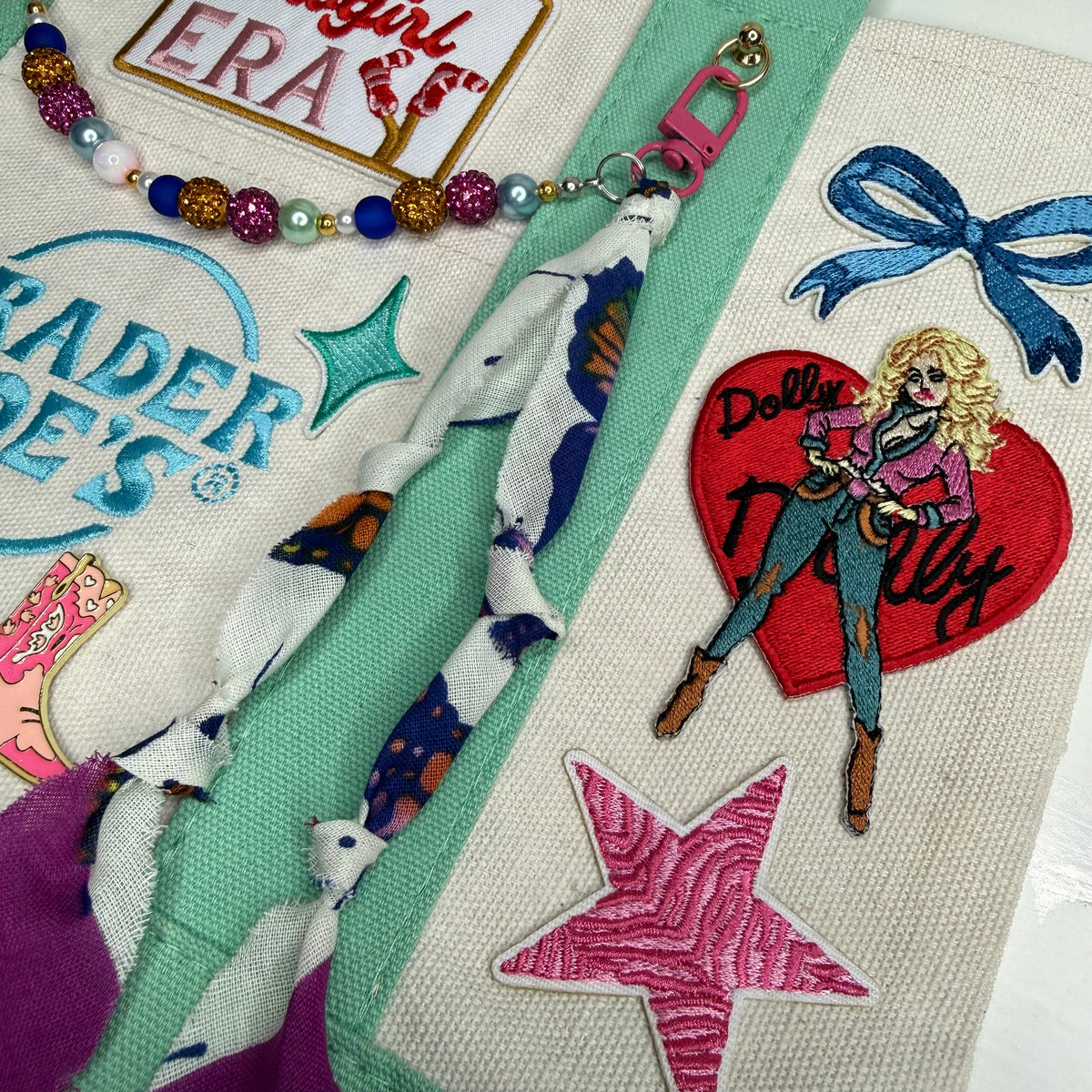 Close-up of the mint and cream Trader Joe's mini tote with the Dolly Parton red heart patch, a blue bow patch, a pink zebra-stripe star, a rhinestone bead chain and the embroidered Trader Joe's logo. Dolly Parton Cowgirl Era mini tote by PrettyMUCH.
