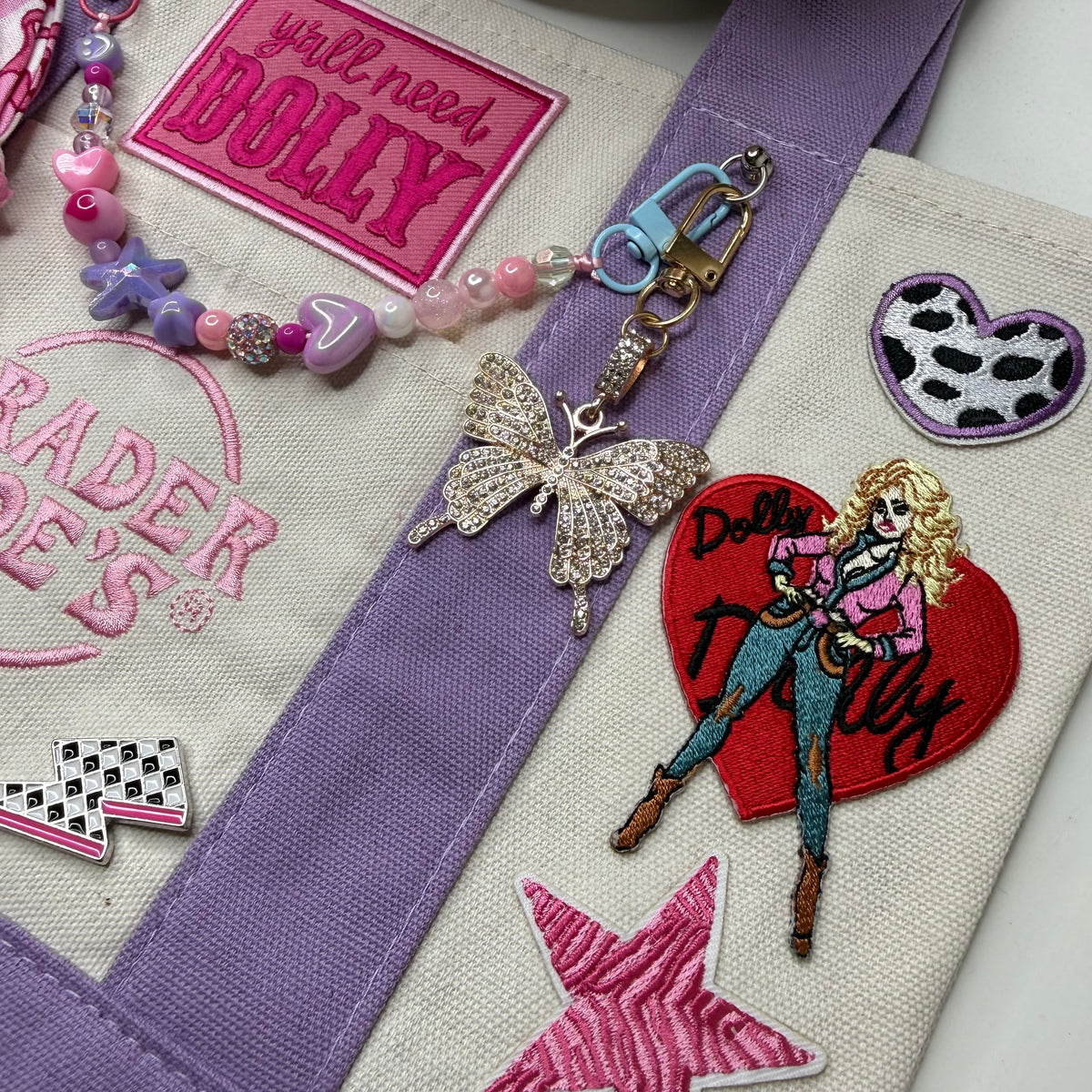 Close-up of the lavender and cream Trader Joe's mini tote with the hot pink 'y'all need DOLLY' patch, a butterfly charm, the Dolly Parton red heart patch and a pink zebra-stripe star patch. Dolly Parton Pretty in Pastel mini tote by PrettyMUCH.