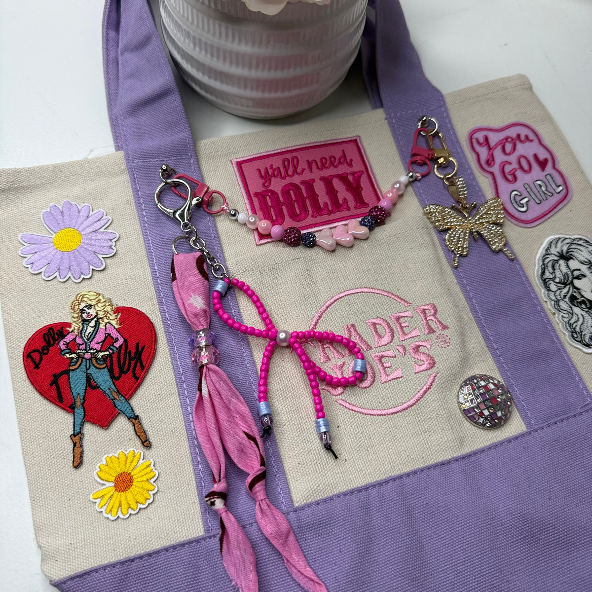 A lavender and cream Trader Joe's mini tote decorated with Dolly patches, daisy patches, a pink beaded bag chain and a butterfly charm, displayed on a white surface. Dolly Parton You Go Girl mini tote by PrettyMUCH.