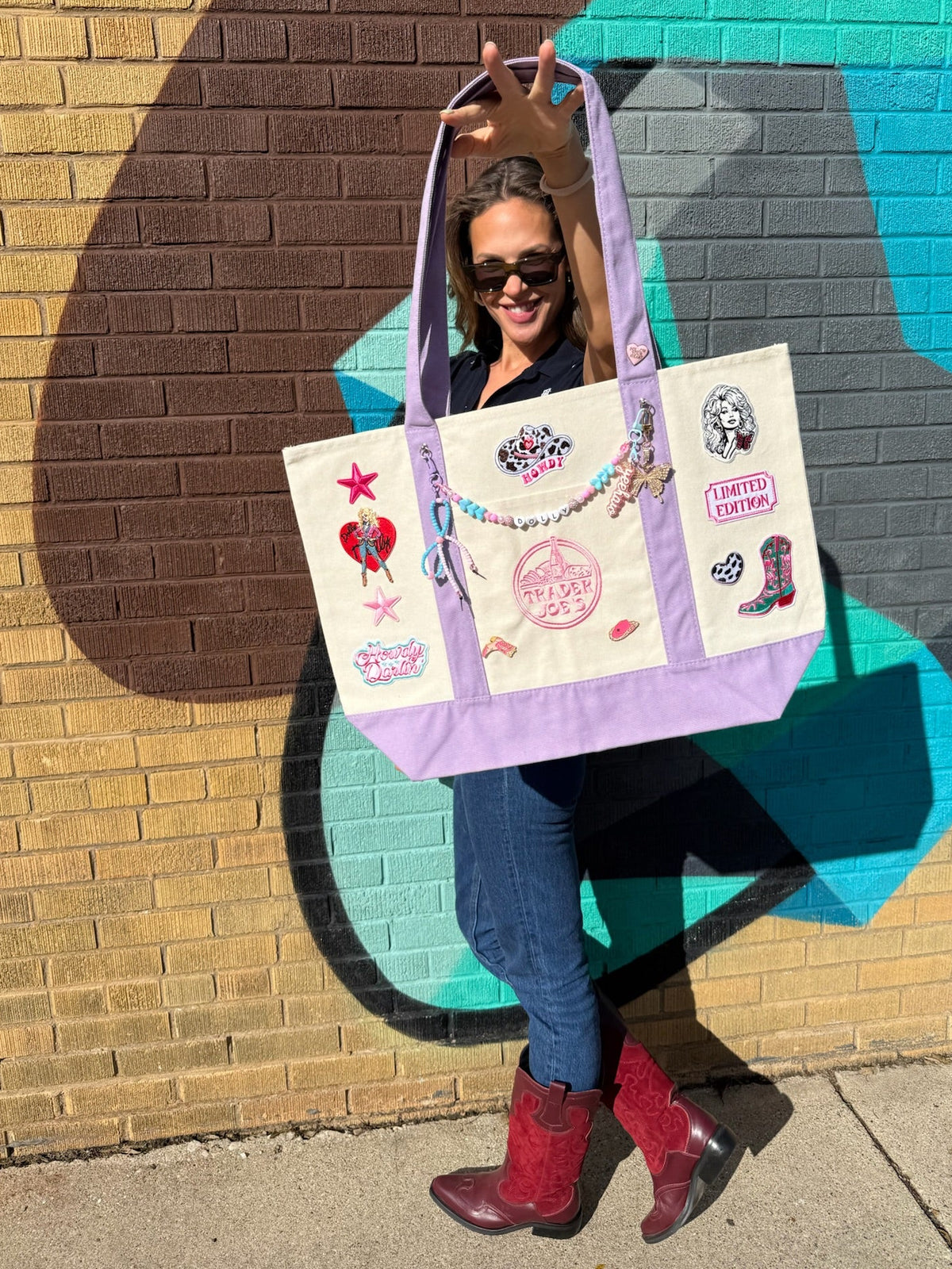 A woman wearing sunglasses, a black top, blue jeans, and red boots holds up a lavender and cream canvas tote bag decorated with various western-themed patches and charms, set against a colorful brick wall with a painted mural. Dolly Parton Howdy Darlin' large tote by PrettyMUCH.