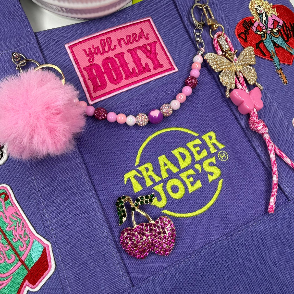 Close-up of the purple Trader Joe's mini tote with the hot pink 'y'all need DOLLY' patch, the neon yellow Trader Joe's logo and a gold cherry heart brooch with pink and green rhinestones. Dolly Parton Boots & Heels mini tote by PrettyMUCH.