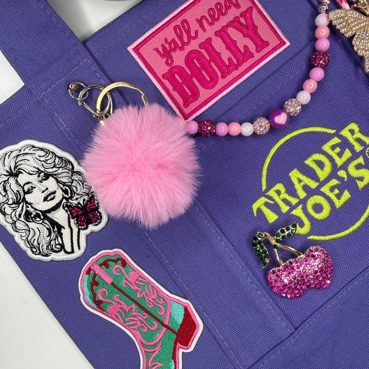 A close-up of a purple fabric tote bag with embroidered patches and accessories, including a pink pom-pom keychain, a pink beaded bracelet, a Trader Joe's patch, and a Dolly patch. Dolly Parton Boots & Heels mini tote by PrettyMUCH.