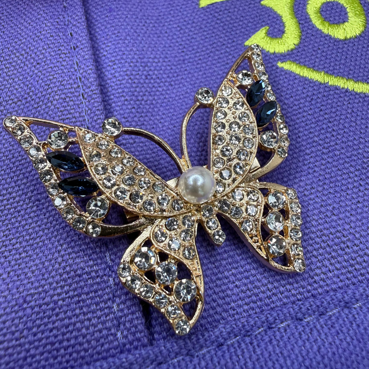 A gold butterfly brooch embellished with clear rhinestones, a central white pearl, and dark blue marquise crystals, resting on a purple textured fabric background. Dolly Parton Aces High mini tote by PrettyMUCH.