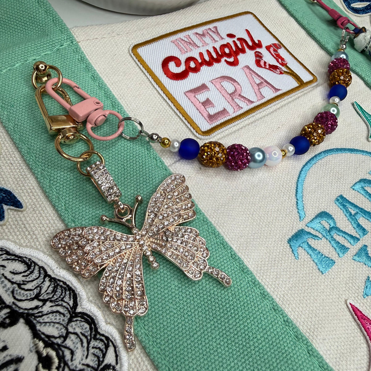 A close-up of a rhinestone-embellished butterfly keychain or bag charm with a pink lobster clasp and a beaded strap, placed on embroidered fabric with a patch reading 'In My Cowgirl Era'. Dolly Parton Cowgirl Era mini tote by PrettyMUCH.