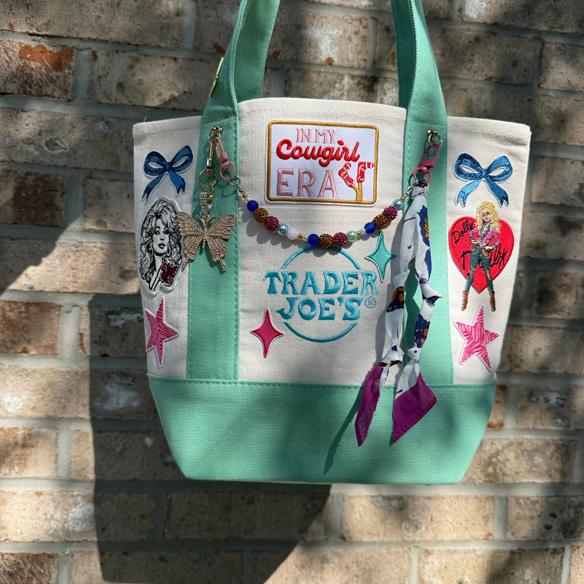 A white canvas tote bag with mint green handles and bottom trim, decorated with colorful embroidered patches, a beaded charm, and a patterned fabric tassel, displayed against a brick wall. Dolly Parton Cowgirl Era mini tote by PrettyMUCH.