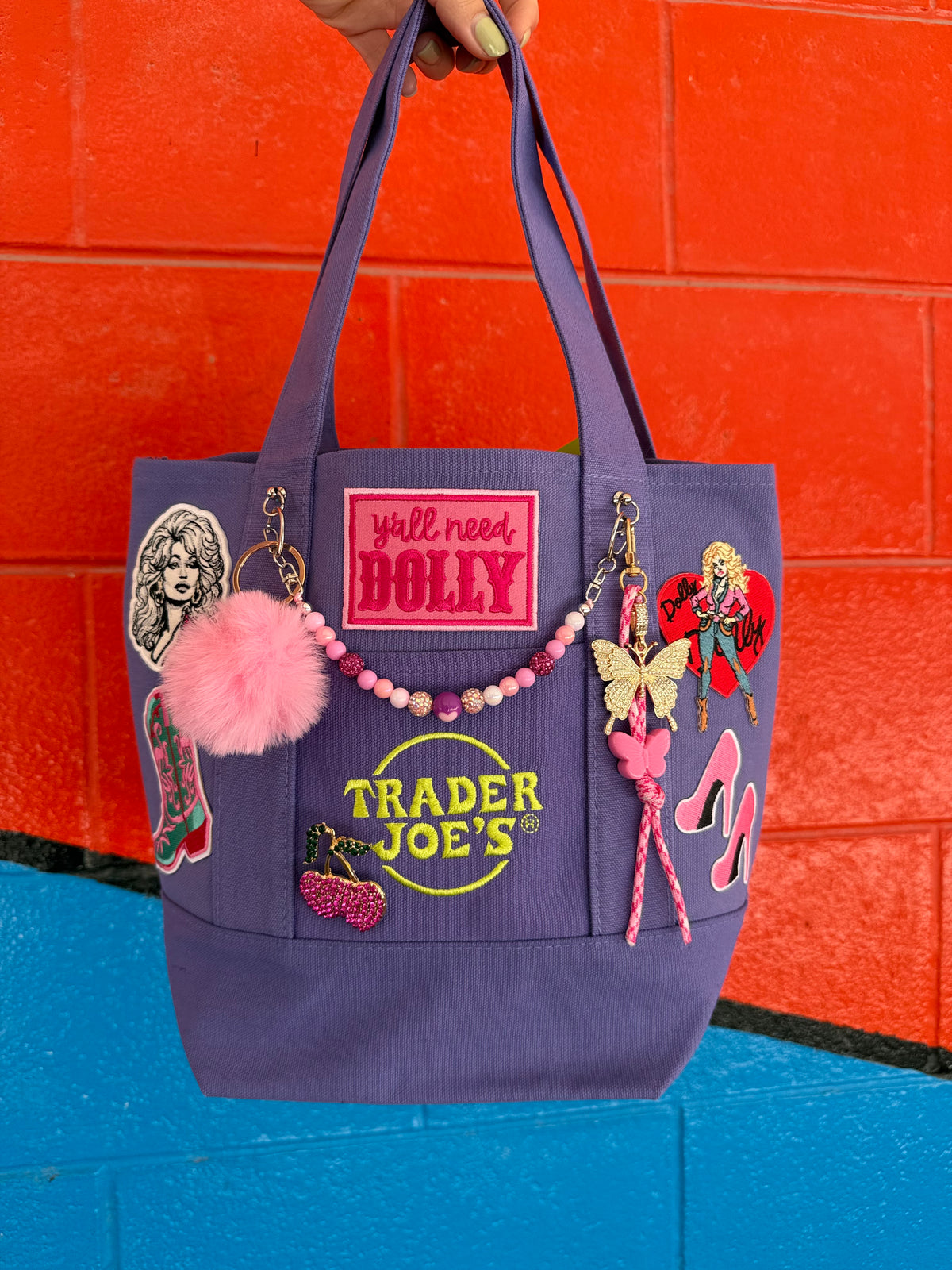 A purple canvas tote bag decorated with various patches, a pink pom-pom, a beaded charm, and a butterfly embellishment, held by a hand against a red and blue brick wall. Dolly Parton Boots & Heels mini tote by PrettyMUCH.