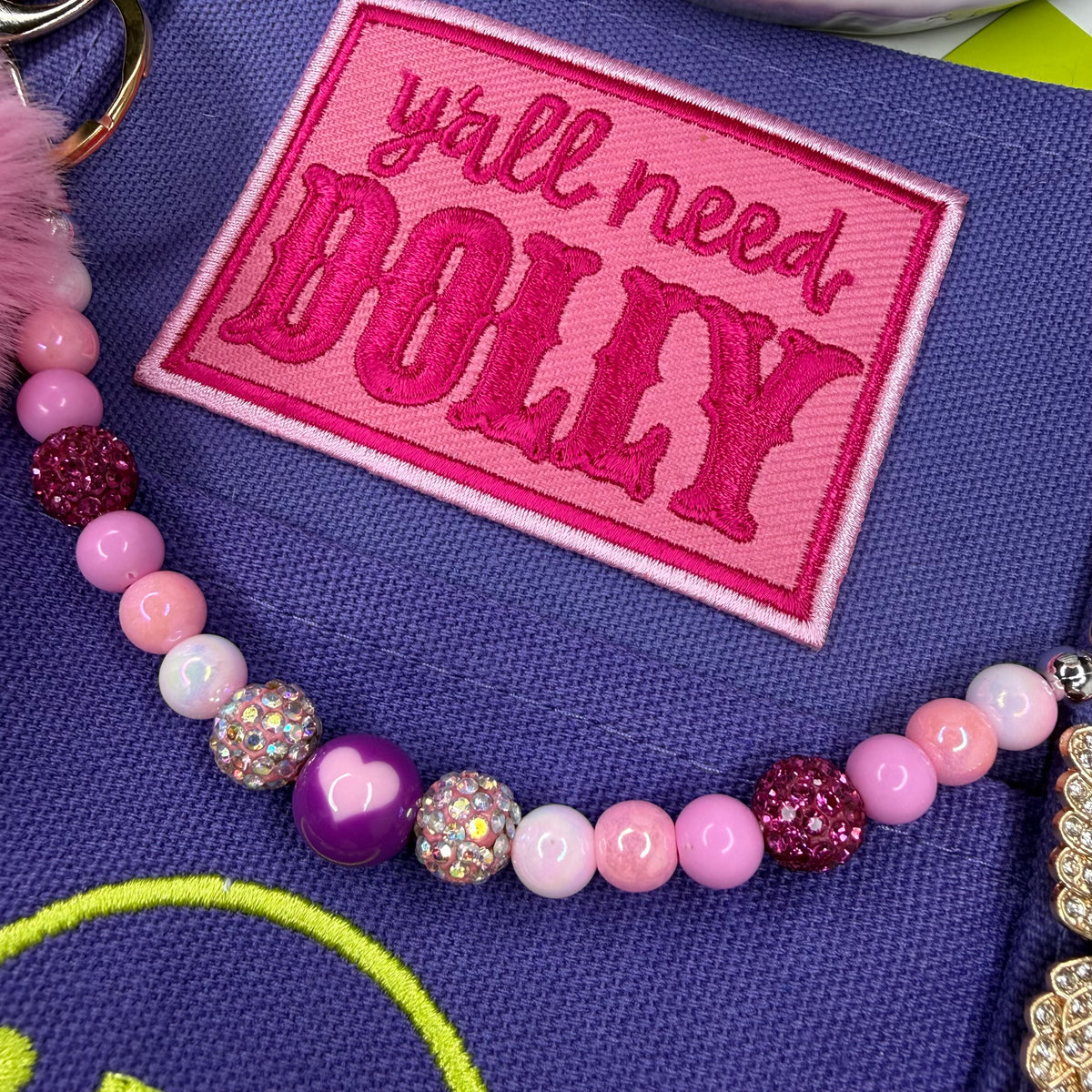 A close-up of a pink embroidered patch reading 'y'all need DOLLY' on a purple fabric item, with a beaded strap or charm featuring pink, purple, white, and glittery beads. Dolly Parton Boots & Heels mini tote by PrettyMUCH.