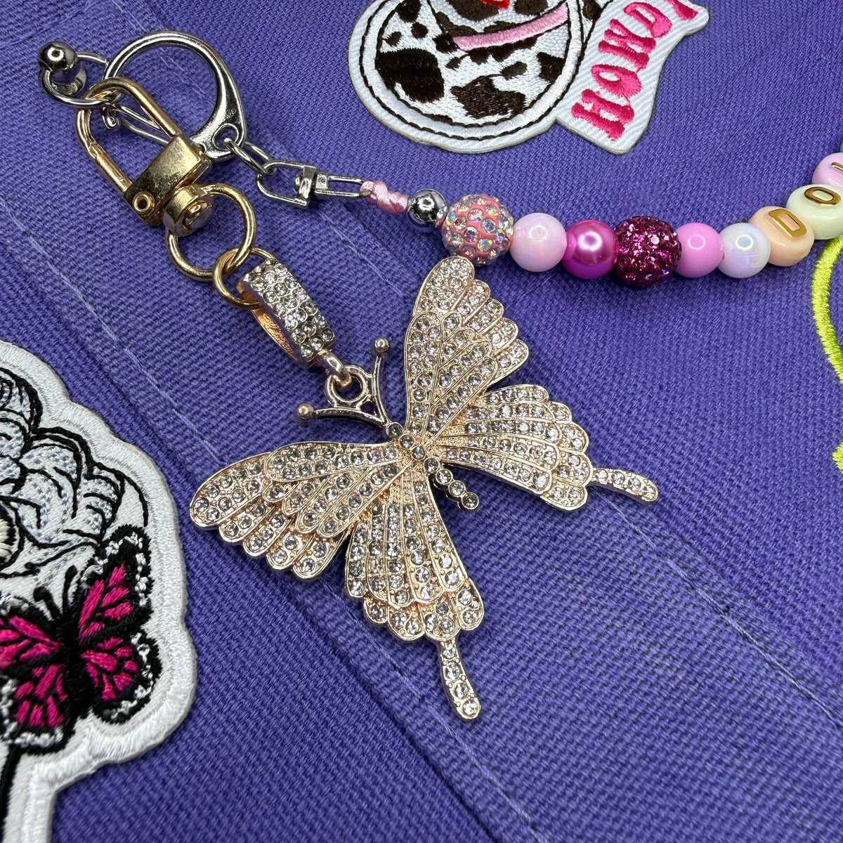 A close-up of a jewelry keychain featuring a butterfly charm covered in clear rhinestones, attached to a gold-tone split ring and lobster clasp, with a strand of pastel and glittery beads on a purple embroidered fabric background. Dolly Parton Sweet but Strong mini tote by PrettyMUCH.