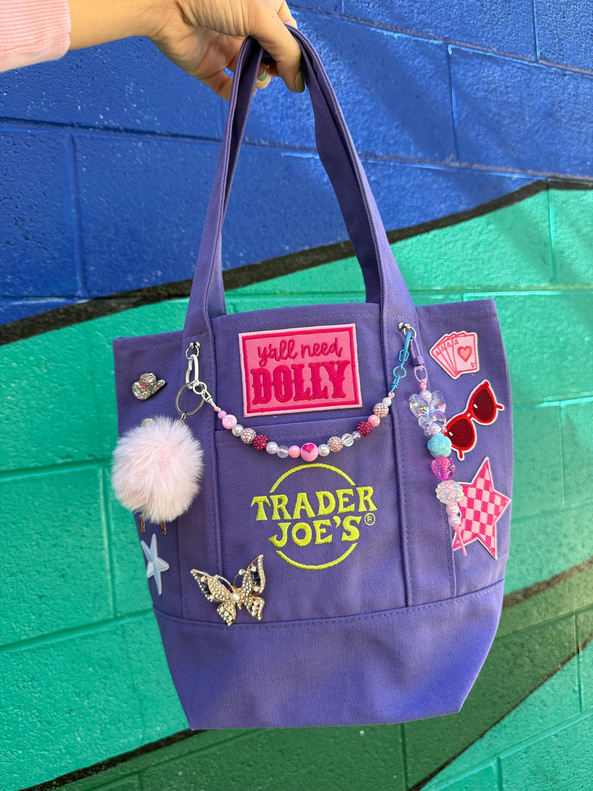 A purple Trader Joe's mini tote with embroidered patches, charms and a pink pom-pom, including the hot pink 'y'all need DOLLY' patch, held by a hand against a blue and green painted brick wall. Dolly Parton Aces High mini tote by PrettyMUCH.